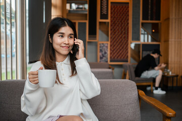 Young woman in a white sweater talking on the phone while holding a coffee cup in a stylish cafe setting.