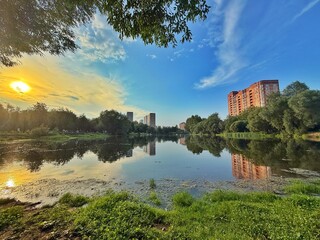 A scenic view of a lake reflects urban buildings and a bright sun under a blue sky. Concept of City nature harmony.