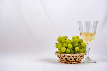 Racimo de uvas verdes en un cestita de mimbré con una copa de vino blanco al lado con fondo blanco 