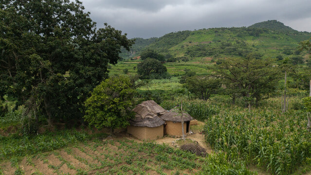 Aerial view of thatched huts nestled amongst verdant fields, backed by rolling hills under a cloudy sky, Jos, Jos North, Nigeria.