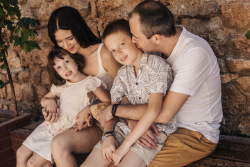 A family of four sits together on a bench. A mother with long dark hair, a father with short hair,...