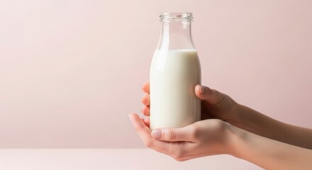 Womans hands holding a bottle of fresh milk against a soft pastel pink background in studio