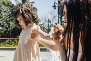 A young Caucasian girl in a light dress holds her mother's hand while holding a teddy bear. The...