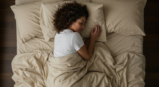 Overhead view of a woman with curly brown hair asleep on her side in a beige bed with a beige duvet and pillows; peaceful slumber, serene expression