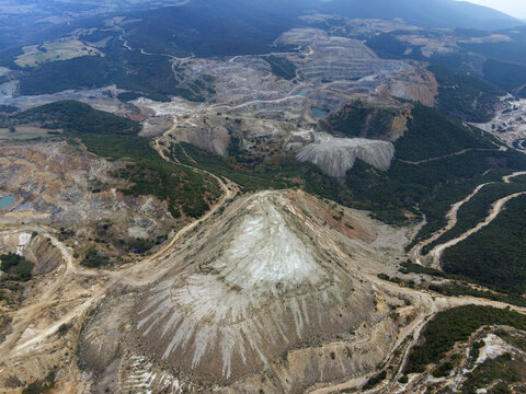 Aerial view of the stark, scarred landscape of a vast mining operation contrasts sharply with the verdant forests in the distance, Vavdos, Greece.