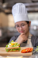Thoughtful Young Asian Female Chef Evaluating Sandwich in Commercial Kitchen