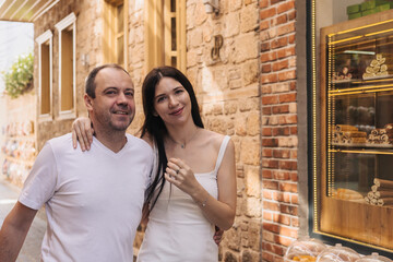 A happy couple stands together in a charming street. The man has short brown hair and a light...