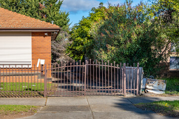 Old metal gate and neglected driveway of a weathered suburban brick house in Australia with rusting fence, overgrown bushes,discarded furniture. a rundown, shabby aging residential neighborhood