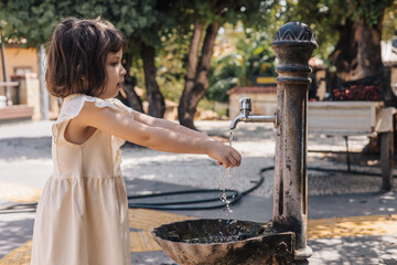 A young girl with short brown hair washes her hands at a water fountain. She wears a light dress and smiles while enjoying the outdoor setting.