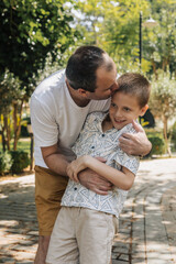 A father hugs his young son in a park. The father is Caucasian with short hair, wearing a white shirt and shorts. The son has short brown hair and smiles happily.