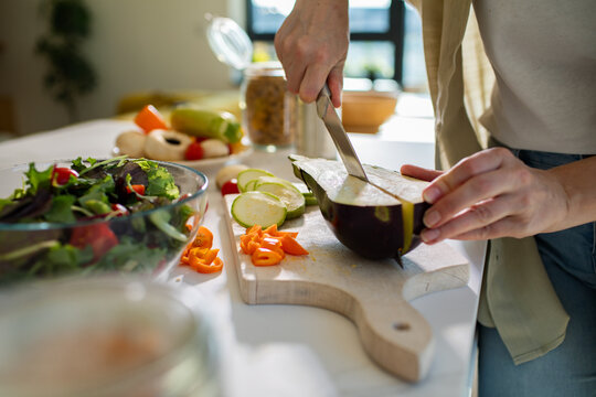 Woman slicing eggplant and vegetables on cutting board for fresh salad in sunny kitchen