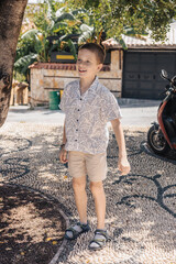 A young Caucasian boy stands outdoors, smiling. He wears a short-sleeved shirt and shorts, with sandals. The background features greenery and a parked scooter.