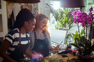 Two smiling female florists using digital tablet in bright flower shop workspace