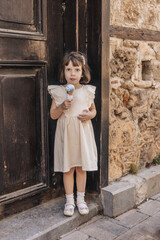A young girl stands by a wooden door, holding an ice cream cone. She has brown hair and wears a light dress with ruffled sleeves. The background features a stone wall.