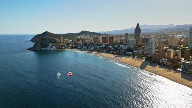 Aerial drone view of the buildings along the coastline with people parasailing in the sea in Benidorm, Spain in daylight
