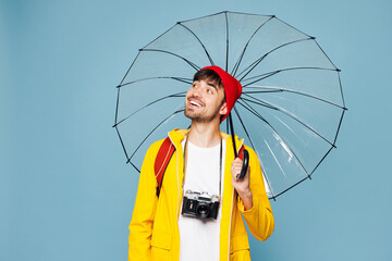 Traveler man wearing hat yellow raincoat casual clothes hold umbrella isolated on plain pastel blue background. Tourist travel abroad in free spare time rest getaway. Air flight trip journey concept.