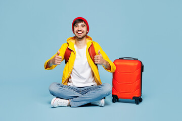 Traveler man wear hat yellow casual clothes sit near bag show thumb up isolated on plain pastel blue background. Tourist travel abroad in free spare time rest getaway. Air flight trip journey concept.