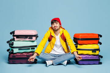 Traveler sad man wear hat yellow casual clothes sit near stack of bags isolated on plain pastel blue background. Tourist travel abroad in free spare time rest getaway. Air flight trip journey concept.