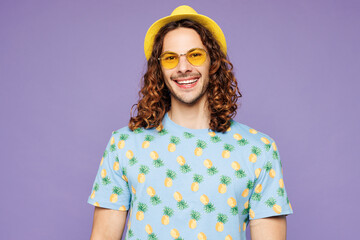 Young smiling happy cheerful fun student man wears blue beach t-shirt yellow hat sunglasses rest near hotel pool look camera isolated on plain purple background. Summer vacation sea sun tan concept.