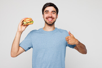 Young smiling happy fun man he wearing blue t-shirt casual clothes hold eat burger show thumb up...