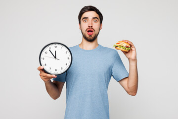 Young shocked surprised man he wear blue t-shirt casual clothes hold in hand clock show time eat...