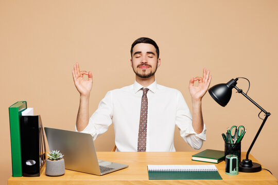 Young employee business man wear white shirt tie sit work at office desk with pc laptop hold hands in yoga om aum gesture relax meditate isolated on plain beige background. Achievement career concept.