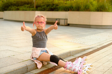 Happy young girl sitting on curb at sunset giving thumbs up, wearing gray tank top, denim shorts,...