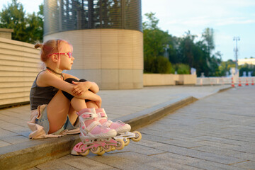 Thoughtful girl in pink glasses sitting on city curb at sunset, her arms wrapped around her knees,...