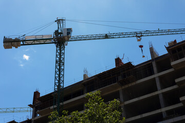 Green tower crane lifting materials above a multi-story concrete building under construction, set against a bright blue sky.