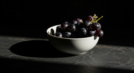 A white bowl filled with dark purple grapes sits on a textured , illuminated by a dramatic light casting deep shadows.