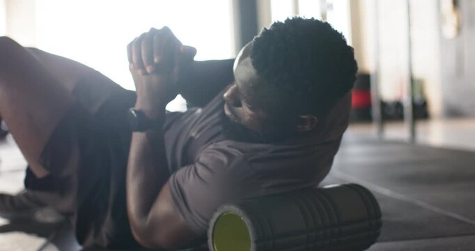 African American man placing foam roller under torso on mat in gym, releasing muscle tension