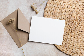 Blank invitation or greeting card mock up with craft envelope and dried flowers on a woven placemat, copy space for creative designs