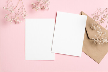 Blank wedding invitation mock up with two white cards and a brown envelope, surrounded by delicate flowers on a soft pink background, ideal for creative design projects