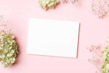 Blank white card mock up surrounded by delicate green and pink flowers on a soft pink background, perfect for wedding invitations or greeting cards with copy space