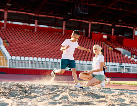 Children running on track at the stadium, jumping a long jump in sand, practice during sports class in school training with coach teacher or referee, little fit boys and girls in sportswear training a