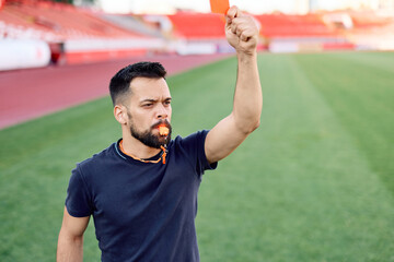 Portrait of a soccer referee blowing a whistle and showing a yellow red card on stadium soccer field