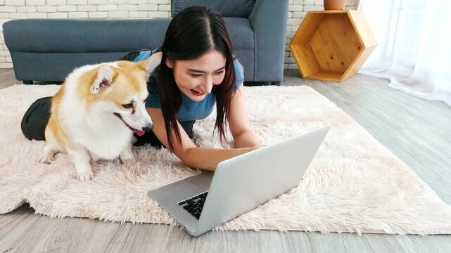 Young woman working on a laptop while lying on a rug beside her Corgi dog at home. A cozy, pet-friendly remote work setup showcasing happiness, productivity, and the comfort of working with pets.