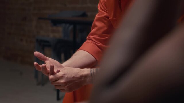 Tilting close-up of face and hands of nervous tattooed Caucasian male inmate in orange uniform, serving prison sentence for gang, drug crimes, fidgeting while reflecting on past life at support group