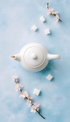 Top view of a white teapot with lid, sugar cubes & peach blossoms on blue smooth surface