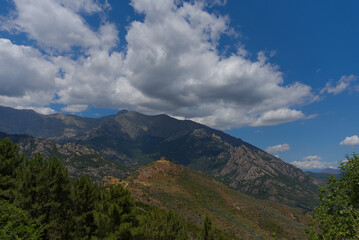 The weathered stone ruins of a small fortress or castle stand on a rocky, brown hill against a backdrop of a vast, forested mountain range under a cloudy sky.