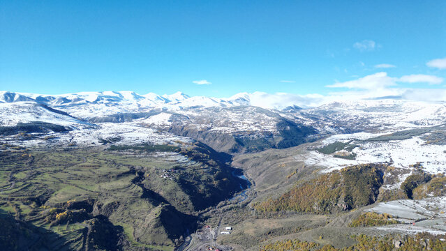 Aerial view of a winding river carves through a valley, flanked by snow-dusted mountains under a crisp blue sky, Posof, Ardahan, T&Atilde;&frac14;rkiye.