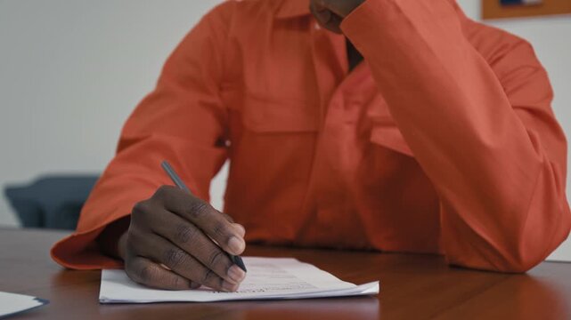 Medium close-up of hand of young African American male prisoner in orange jumpsuit learning to write resume, while participating in rehabilitation program to train in social life skills