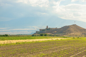 Khor Virap. An ancient Armenian monastery. Stands on a hill overlooking cultivated fields. With Mount Ararat looming in the background under a cloudy sky. Serene and picturesque landscape of Armenia