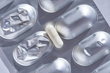 Single white capsule on blister pack surrounded by empty silver foil pill pockets illuminated by...