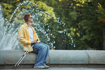 Blind man with assistive trekking poles sitting near urban fountain in casual clothing. Concept of travel, assistive tech, urban space inclusion, and social accessibility.
