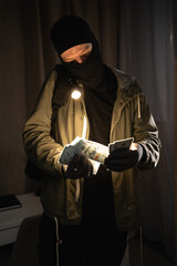Burglar in black balaclava committing burglary counting stolen money in strangers house