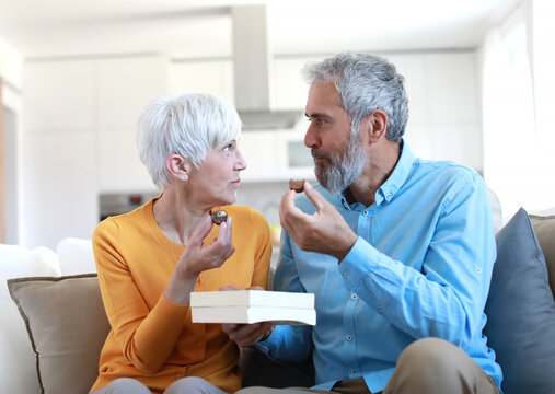 Portrait of a happy senior couple embracing eating chocolate praline from a gift box and having fun at home