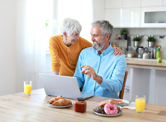 Portrait of an elderly senior couple having breakfast and looking at a laptop at home. Happy healthy affectionate senior couple eating and sitting at kitchen table having fun enjoying morning meal tog
