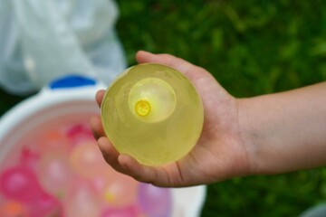 A child holds a yellow water balloon over a grassy area, surrounded by a bucket filled with...
