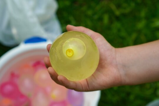 A child holds a yellow water balloon over a grassy area, surrounded by a bucket filled with colorful balloons. The image captures a playful summer activity showcasing joy and anticipation.